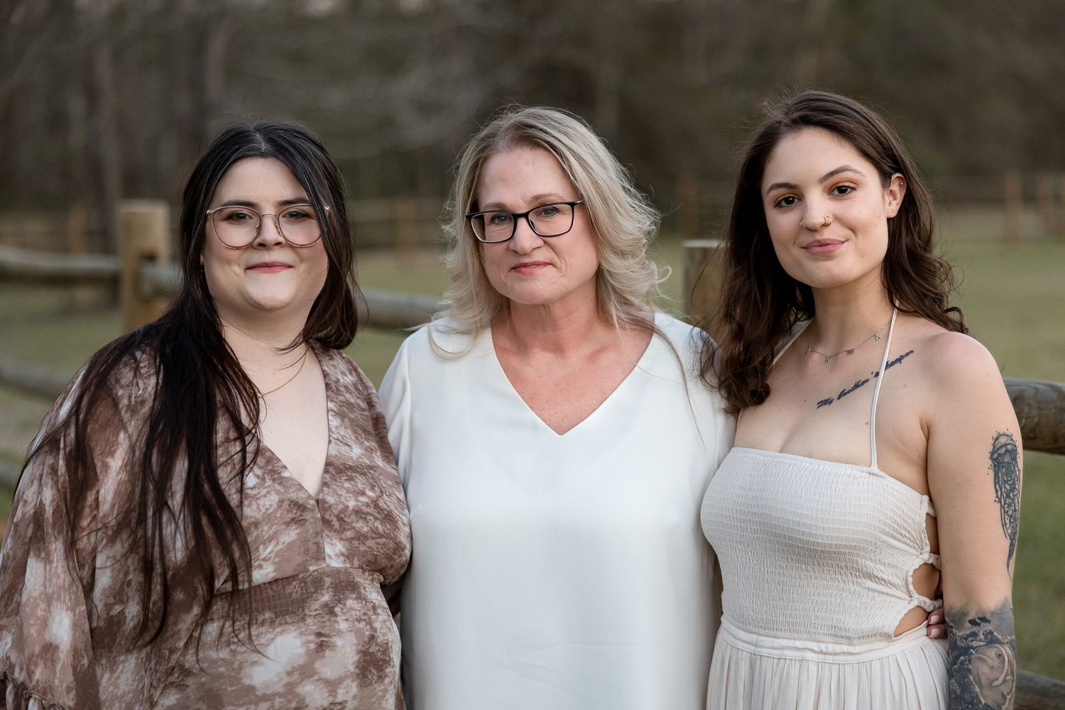 Three women smiling together outdoors in a wooded setting, wearing casual attire for a family portrait.