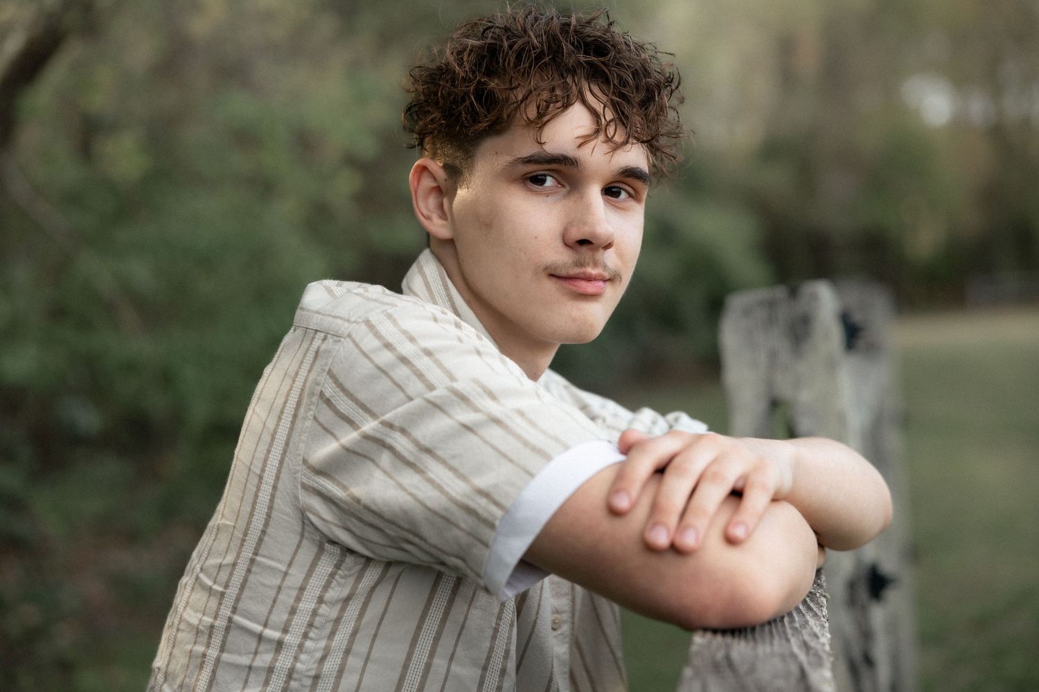 Young man with curly hair leaning on a wooden fence outdoors, wearing a striped linen shirt.