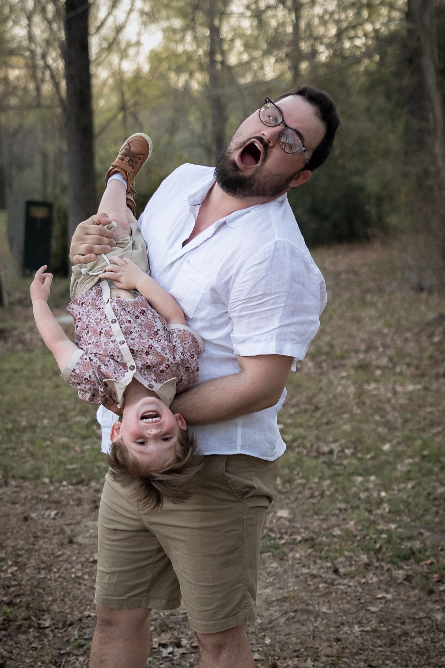 A bearded man with glasses playfully holds a laughing toddler upside down in a wooded outdoor setting.