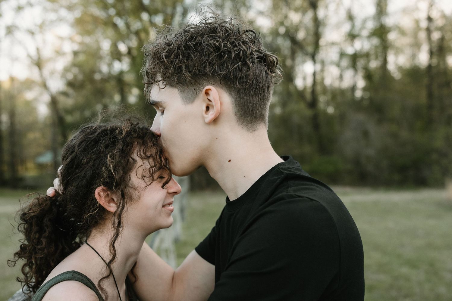 A young couple sharing a tender forehead kiss outdoors in a green park setting.
