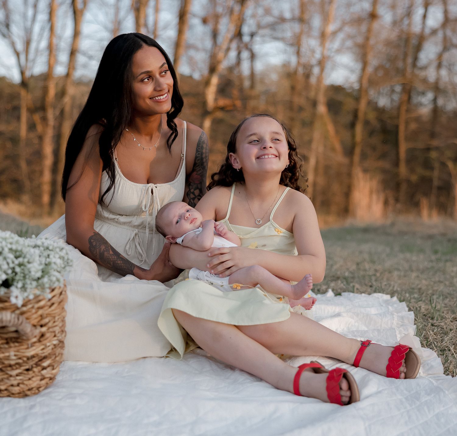 A woman sits on a white blanket outdoors holding a baby beside a smiling young girl in a yellow dress.