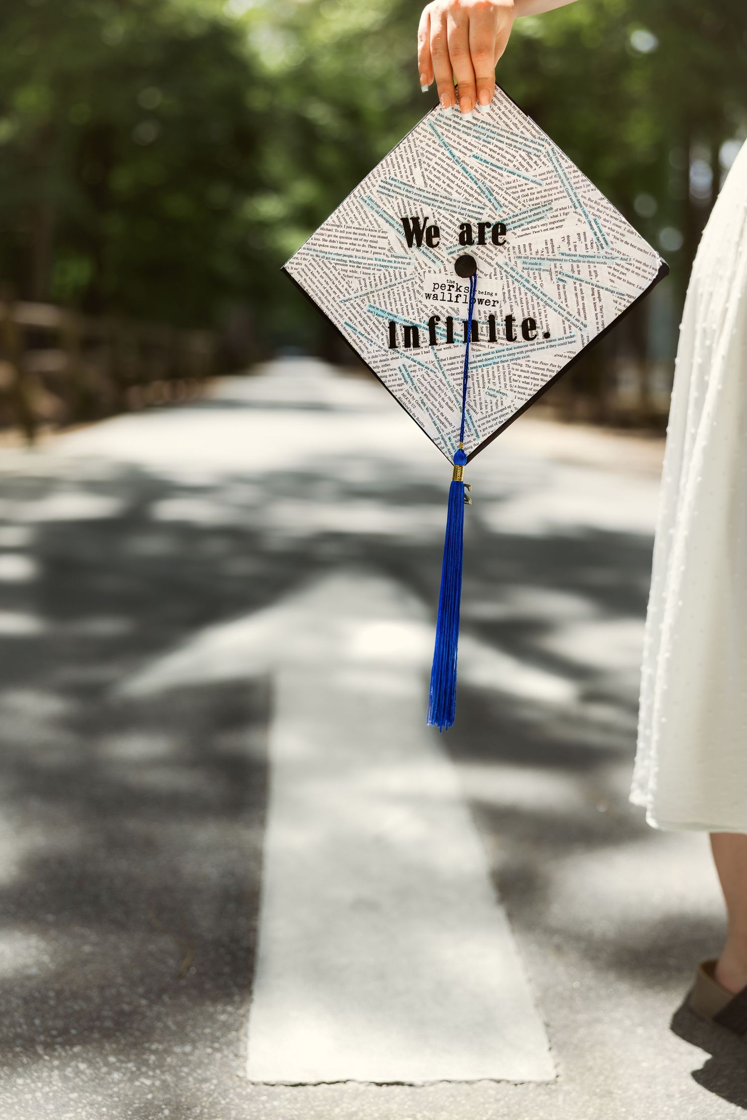 Graduate holding decorated mortarboard cap reading 'We Are Infinite' with blue tassel outdoors.