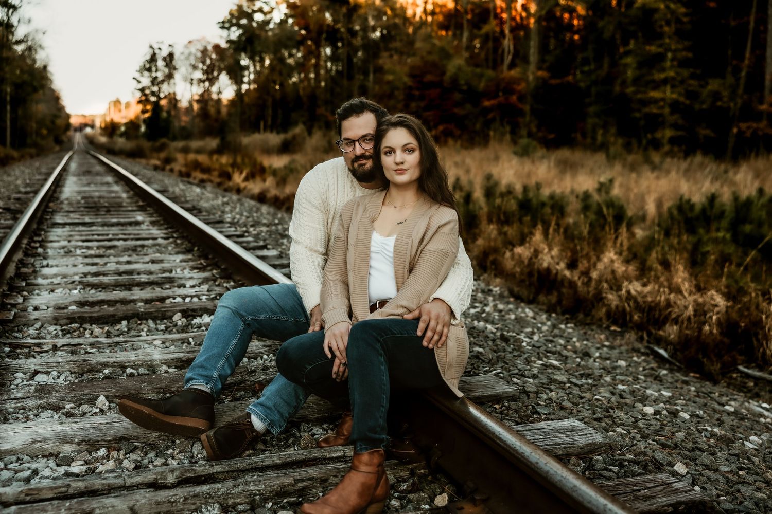 Couple in casual attire posing together on rustic railroad tracks during sunset.