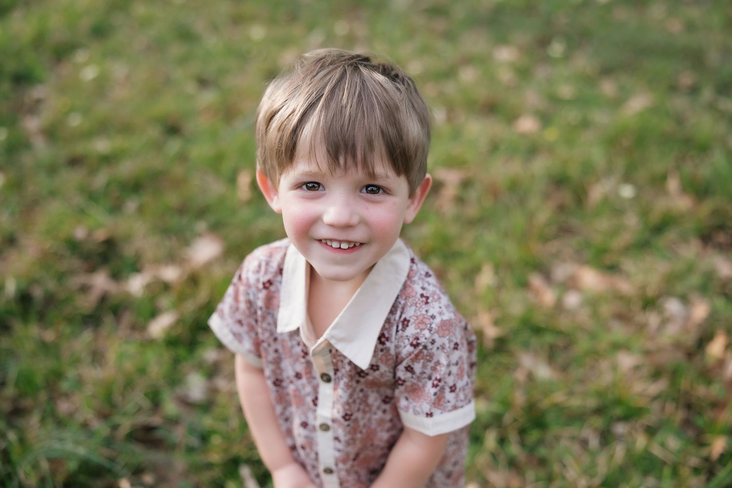 Smiling young child with brown hair wearing a floral collared shirt, posing outdoors on green grass.