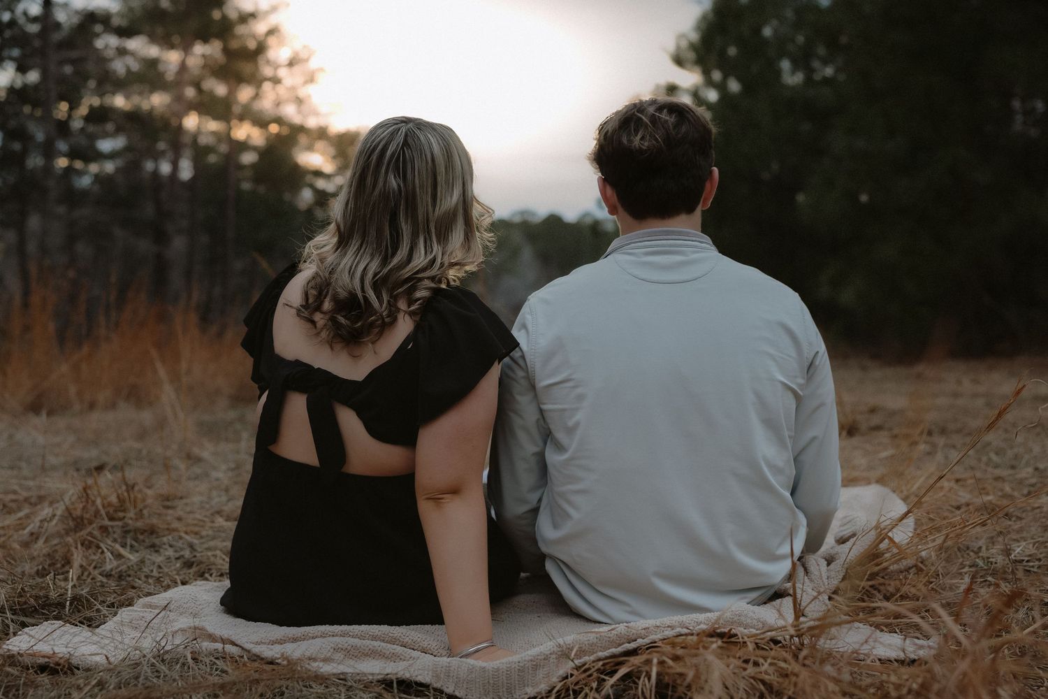Couple sitting together on a blanket in a field at sunset, viewed from behind, surrounded by tall grass and trees.