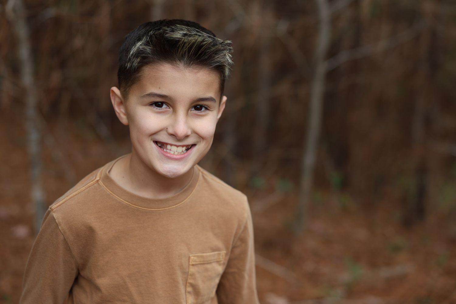 Child in brown shirt smiles warmly during outdoor autumn photoshoot.