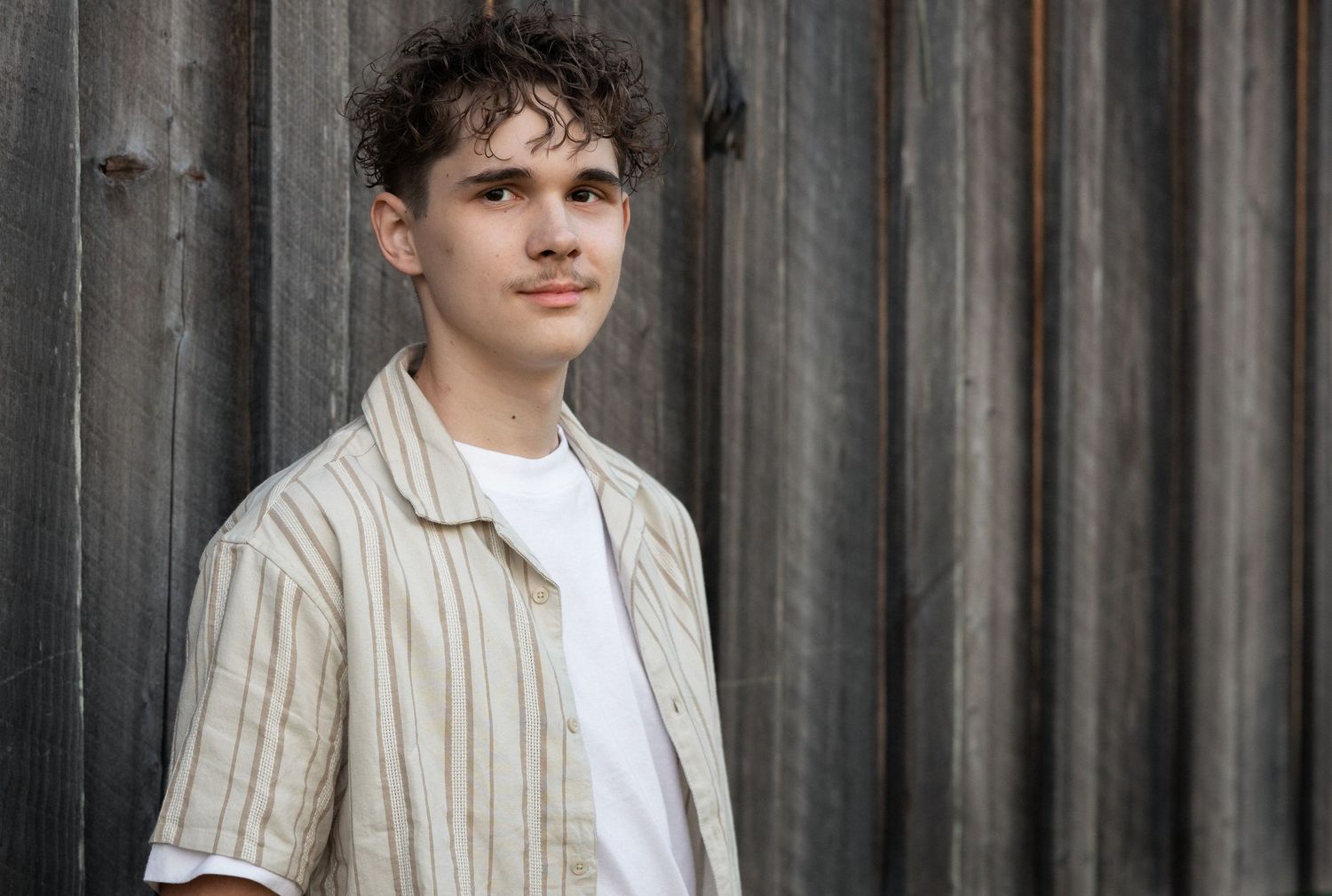Young man with curly hair wearing a striped linen shirt poses against a rustic wooden wall backdrop.