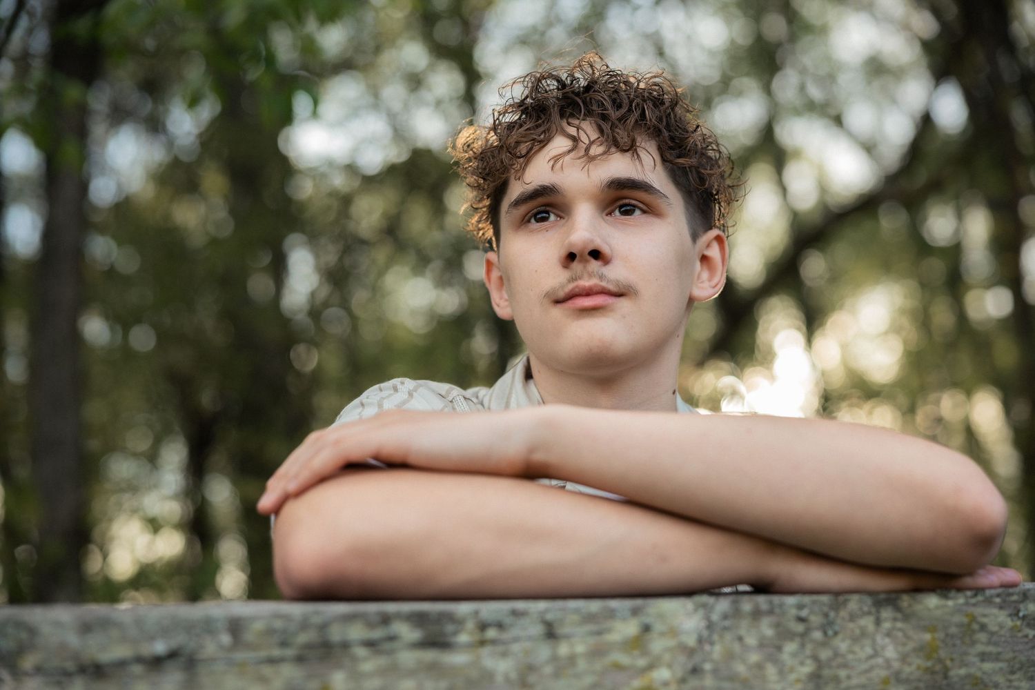 Young man with curly hair resting arms on stone ledge, gazing thoughtfully in a lush green outdoor setting.