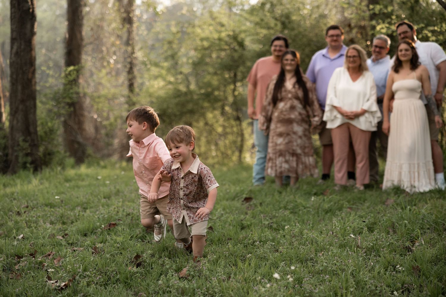 Two young boys run through a grassy field while family members gather in the background during an outdoor photo session.