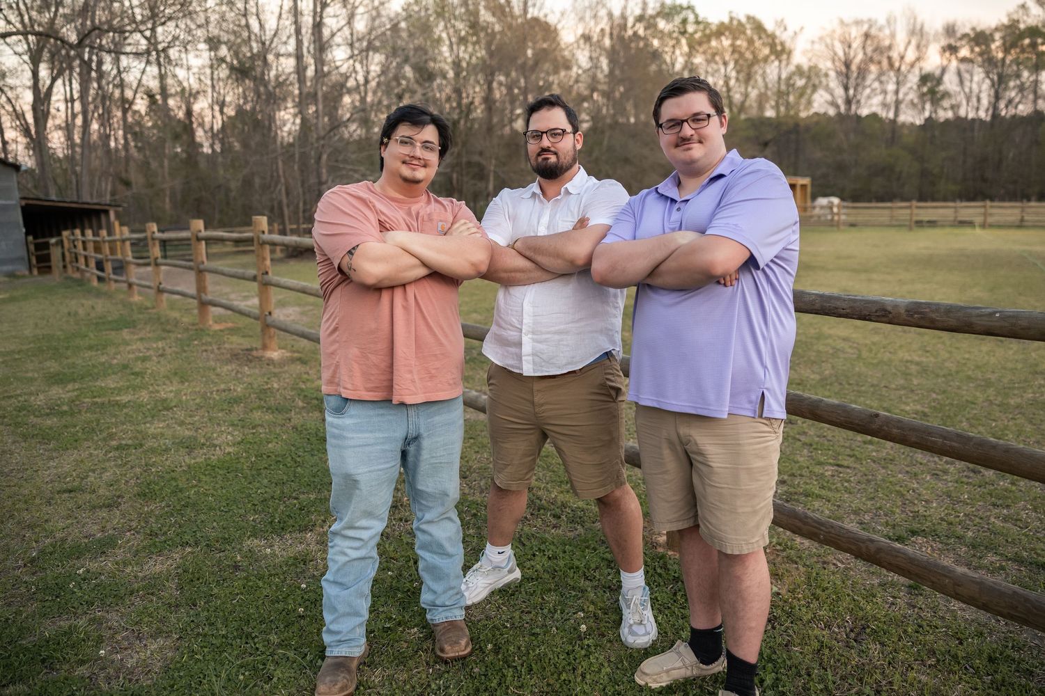 Three men standing with arms crossed by a wooden fence in a rural field at dusk.