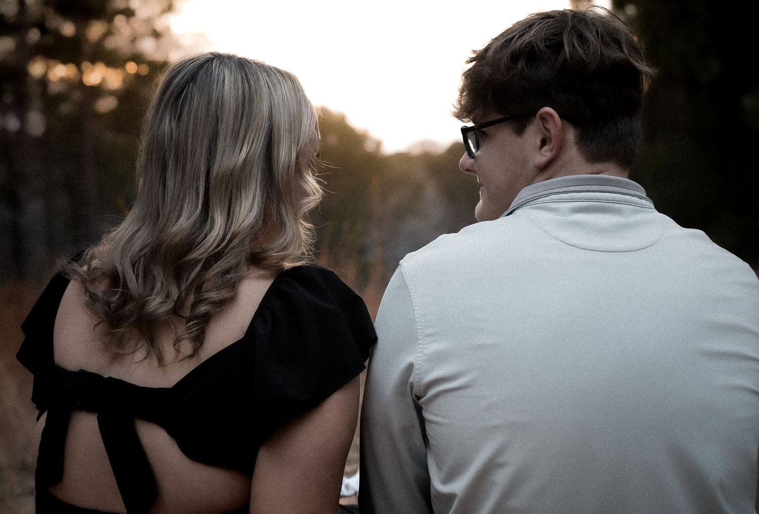 A couple seen from behind sharing a romantic moment outdoors at sunset, woman in black dress beside man in gray shirt.