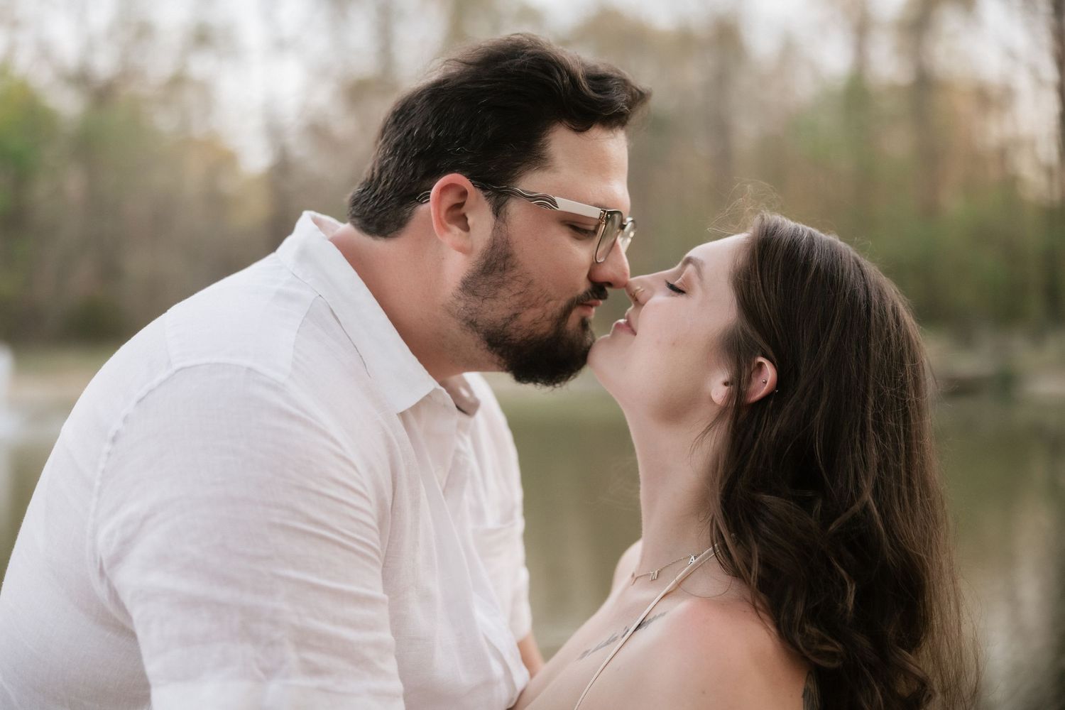 Couple sharing a romantic kiss outdoors near a lake, surrounded by trees in soft natural light.