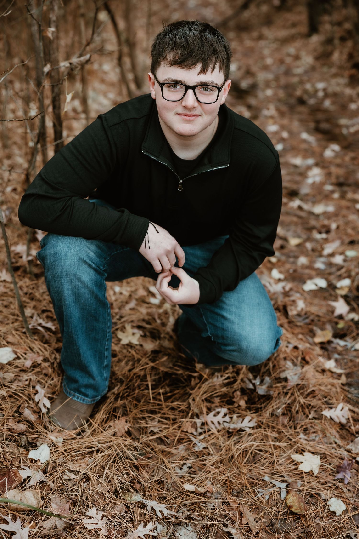 Person in black jacket and jeans crouches to examine fallen autumn leaves in a forest setting.