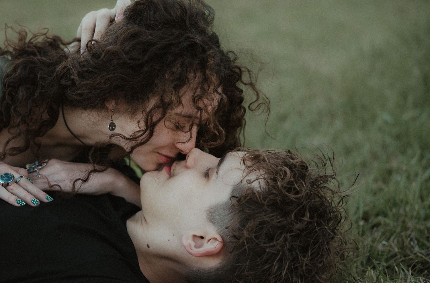A couple with curly hair sharing a romantic kiss while lying on green grass outdoors.