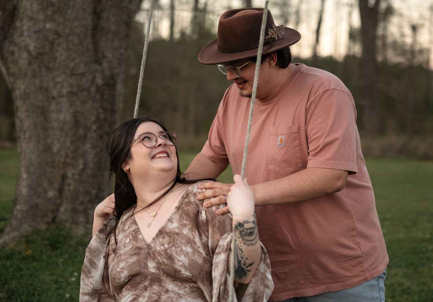 A couple shares a tender moment outdoors, woman in floral top on swing, man in brown hat smiling down at her.