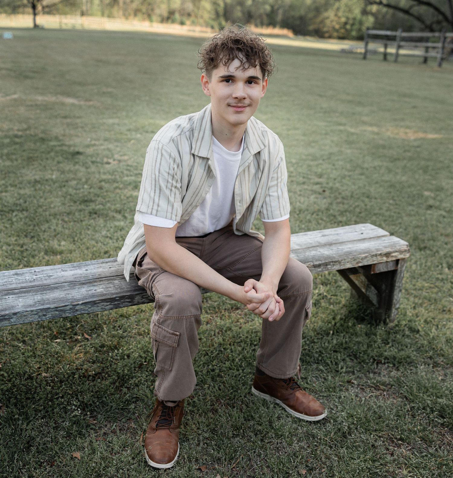 Young man in striped shirt and khaki pants sitting on a rustic wooden bench outdoors on green grass.