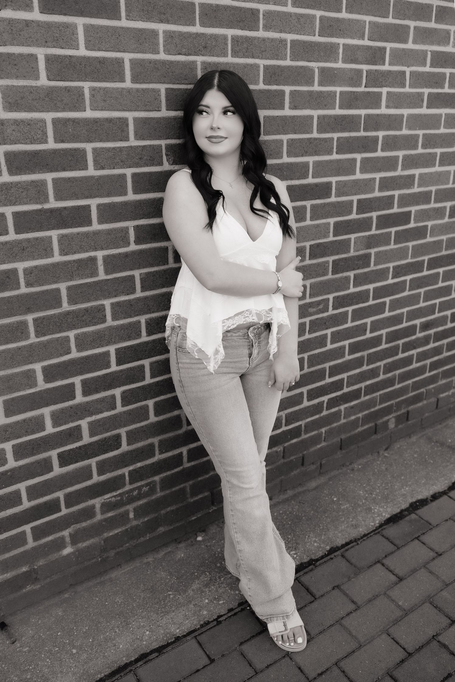Young woman in white top and flare jeans leaning against a brick wall in a black and white portrait photo.