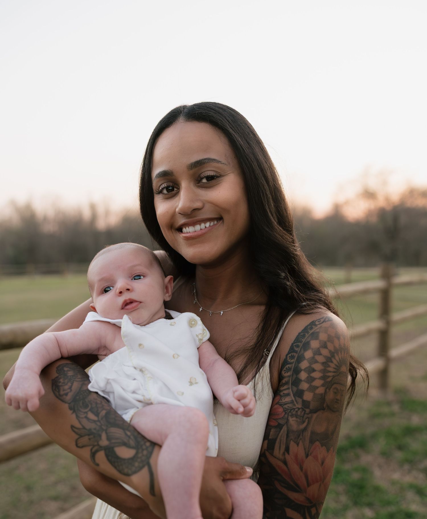 Smiling tattooed woman holding a newborn baby in white outfit outdoors at golden hour near a wooden fence.