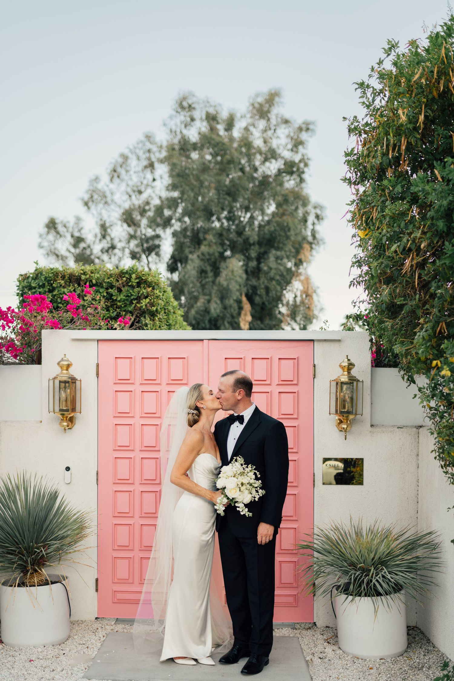 Louis Willard Photo Palm Springs Bride and groom kiss in front of pink doors with bougainvillea at Palm Springs wedding.