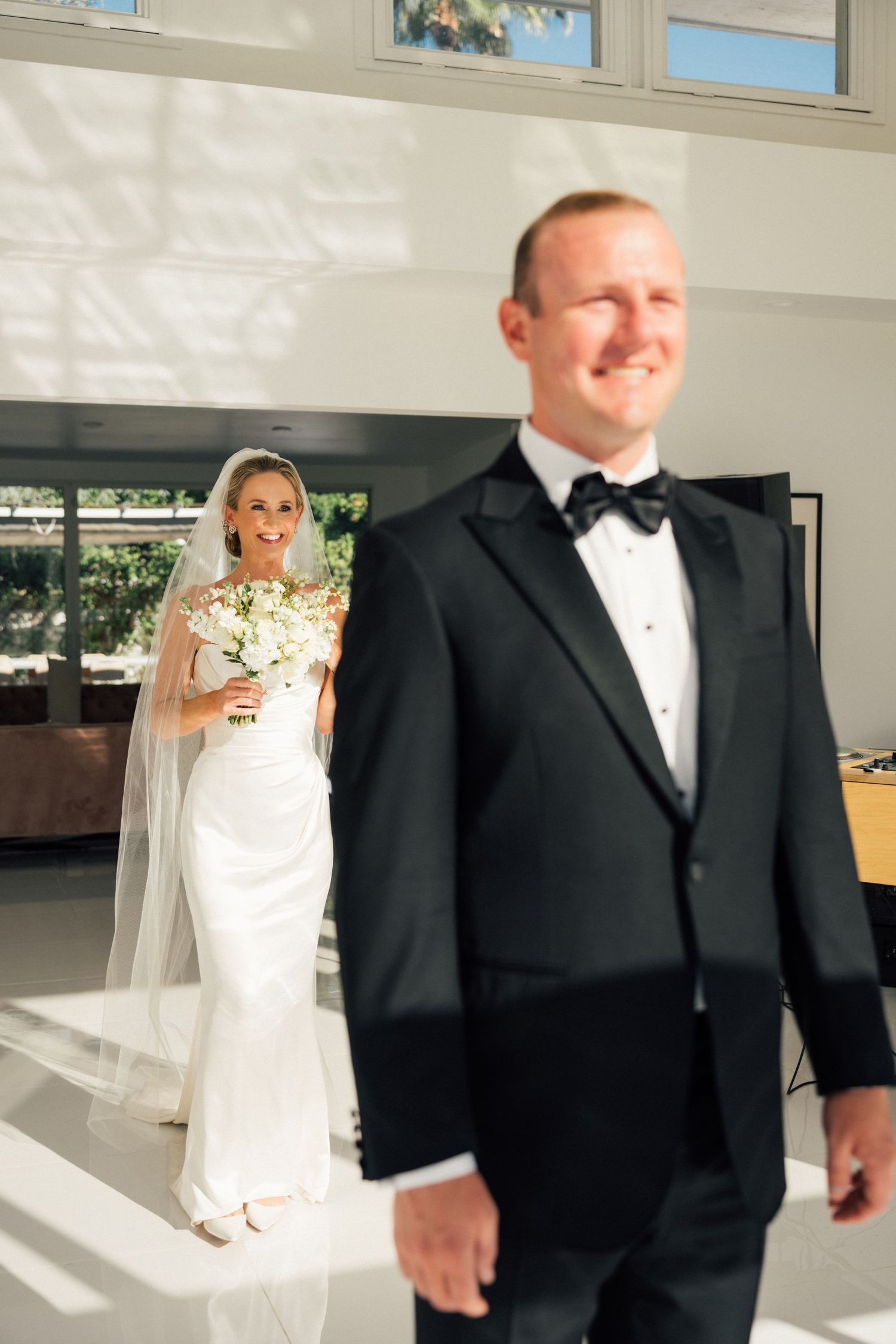 Louis Willard Photo Palm Springs Groom in black tuxedo smiles as bride in white gown and veil approaches for their first look.