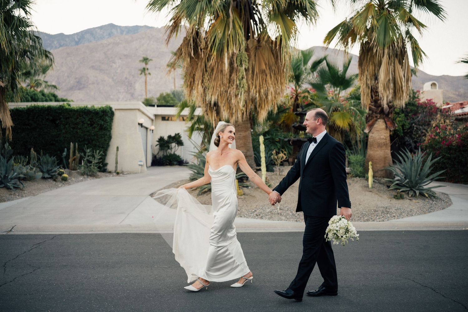 Louis Willard Photo Palm Springs Bride and groom walk hand in hand past palm trees with mountain views in Palm Springs.
