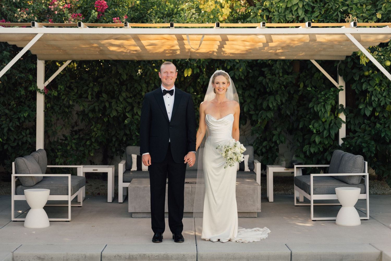 Louis Willard Photo Palm Springs Bride and groom pose together under a pergola surrounded by lush greenery at their outdoor wedding.