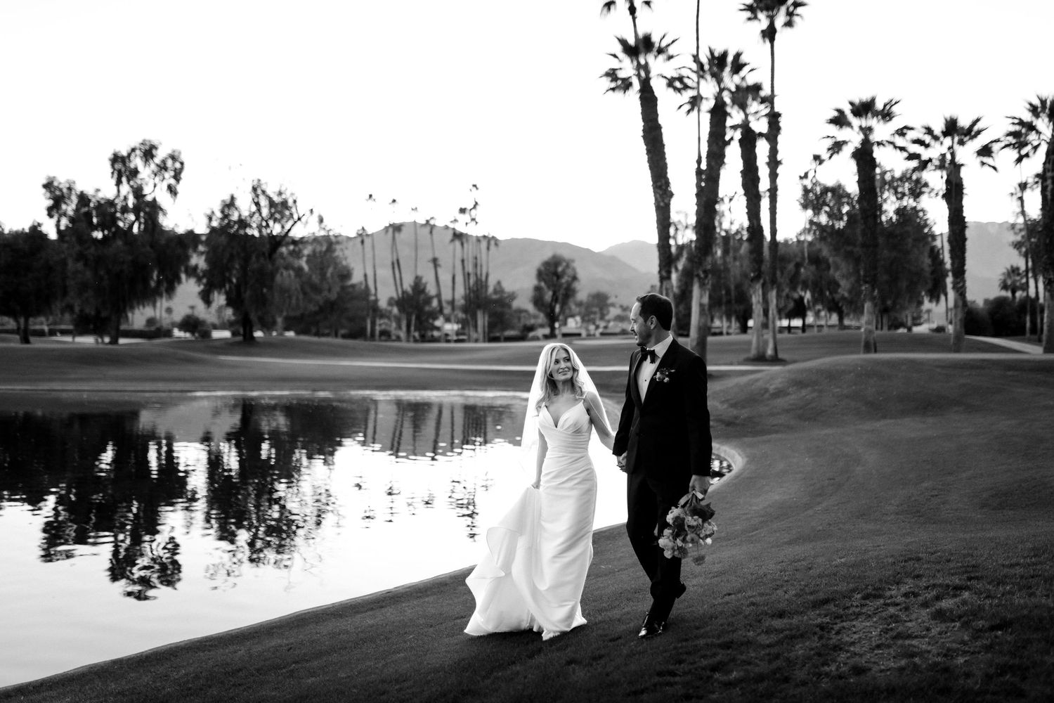 Black and white photo of a bride and groom walking hand in hand by a pond at a golf course wedding venue.