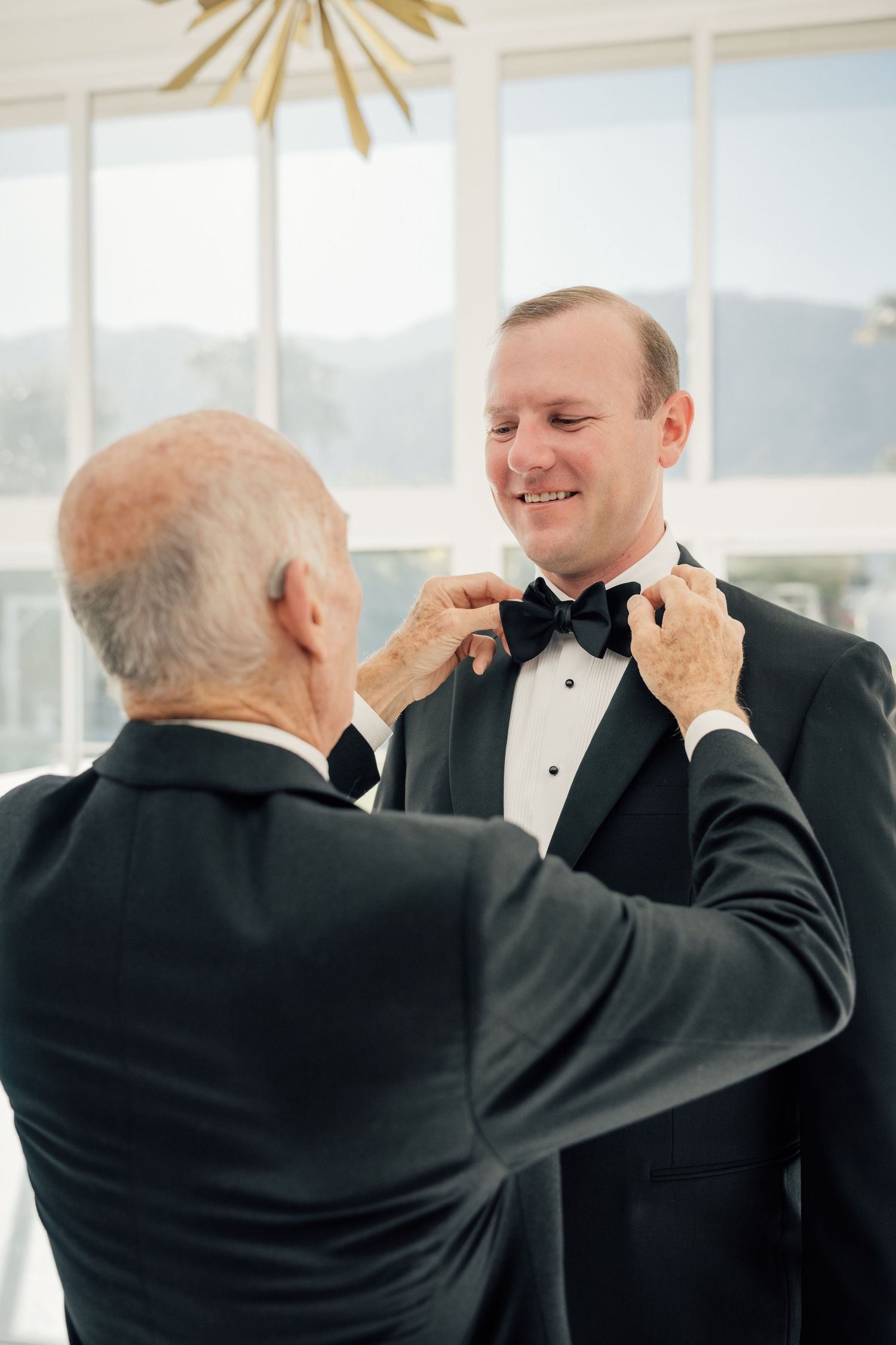 Louis Willard Photo Palm Springs Father adjusts groom's black bow tie as they share a tender moment before the wedding ceremony.