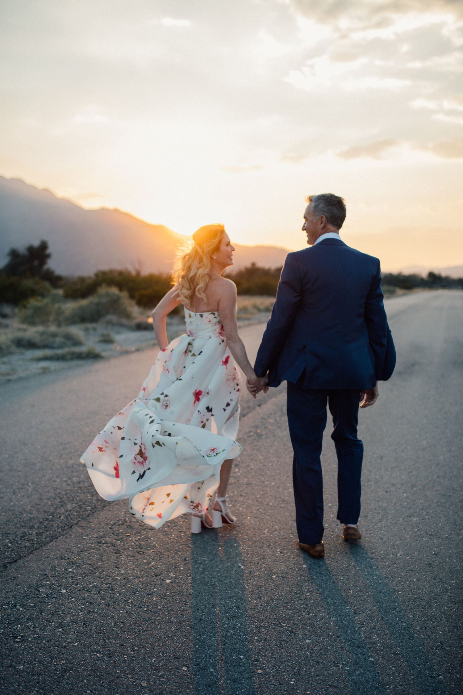 Couple holding hands walking down a desert road at sunset with mountains in the background.