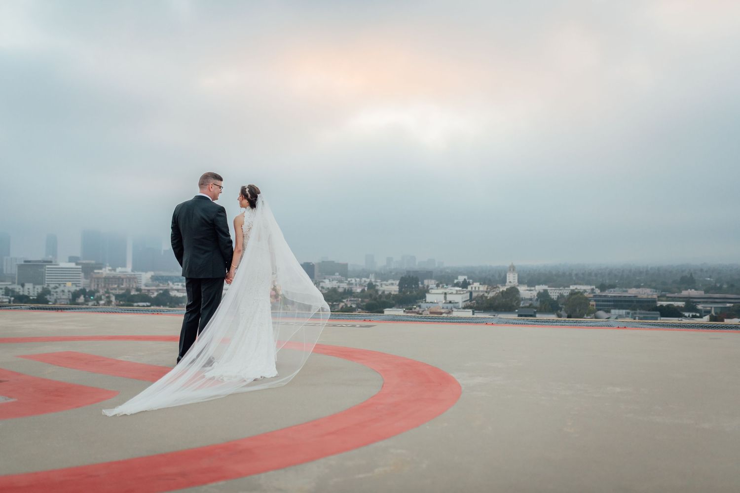 Bride and groom stand on rooftop helipad with city skyline backdrop, bride's long veil trailing behind her.