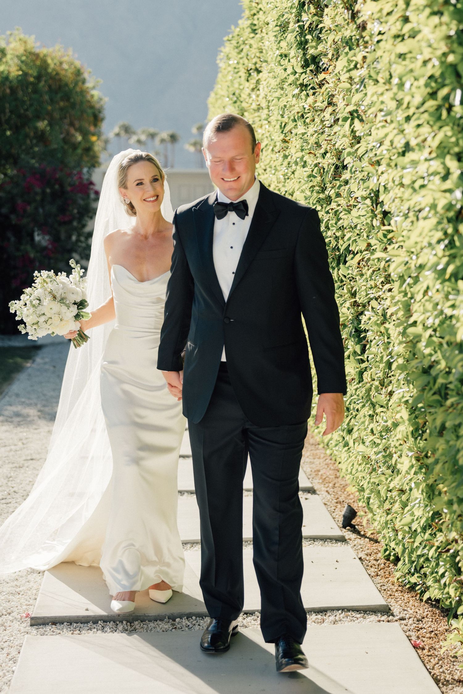 Louis Willard Photo Palm Springs Bride and groom walk hand in hand along a garden path lined with tall green hedges.