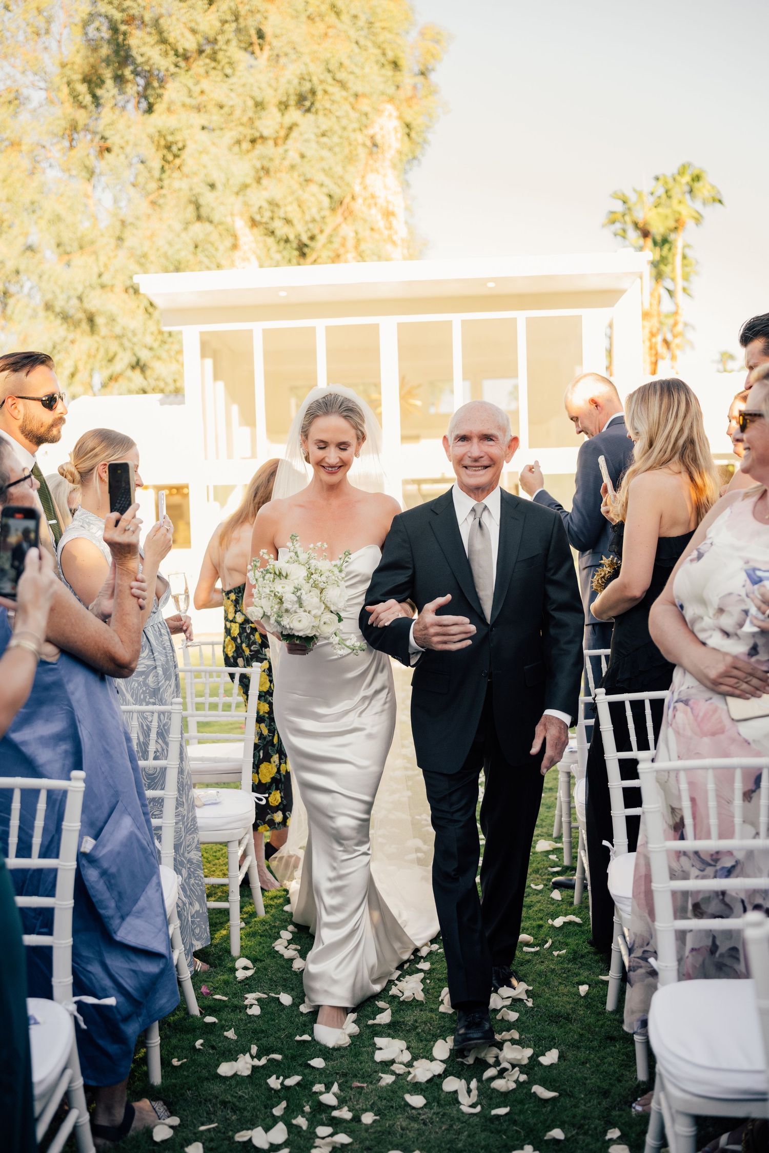 Louis Willard Photo Palm Springs Bride walks down the aisle with escort at outdoor wedding ceremony, rose petals on grass.