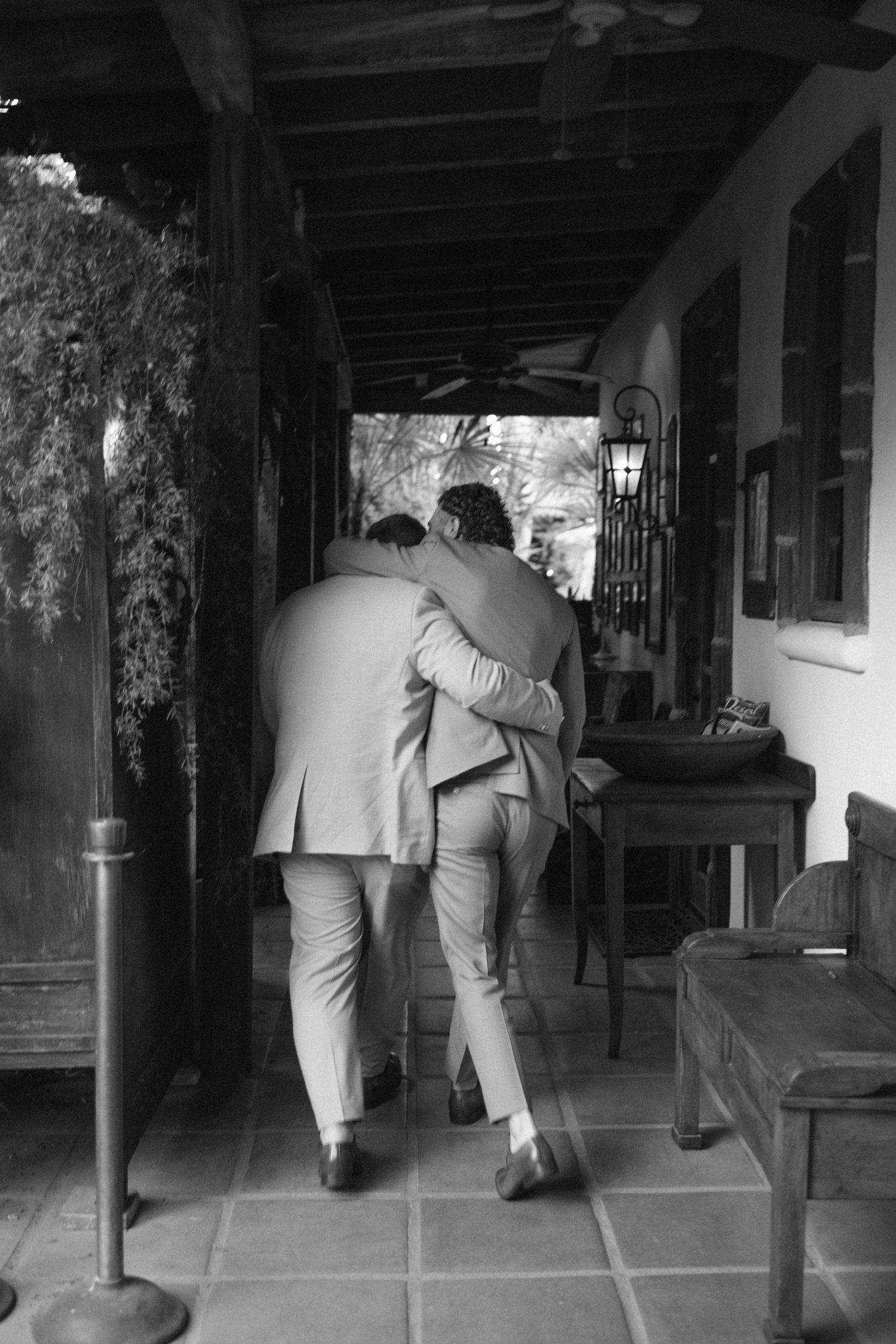 Black and white photo of two grooms embracing while walking down a covered corridor.