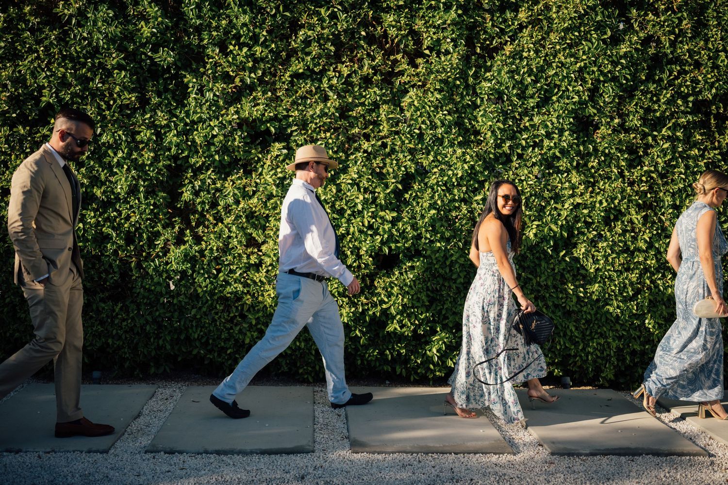 Louis Willard Photo Palm Springs Wedding guests walk along a sidewalk beside a lush green hedge wall on a sunny day.
