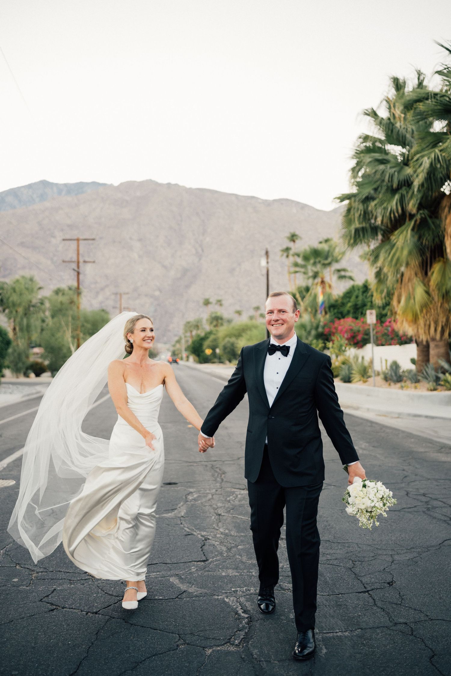 Louis Willard Photo Palm Bride and groom hold hands walking down a sunny Palm Springs street with mountains and palm trees in the background