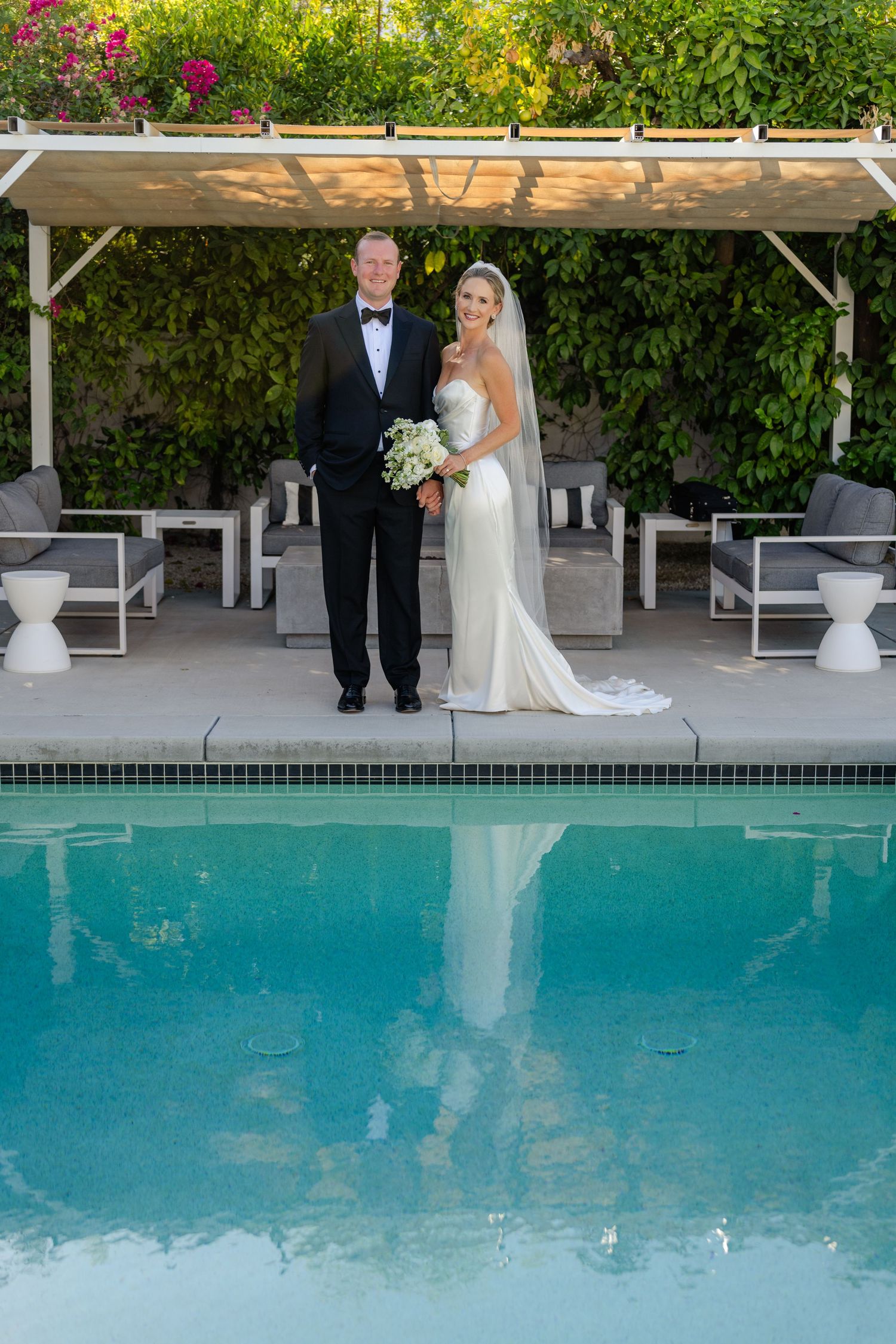 Louis Willard Photo Palm Springs Bride and groom pose elegantly by a pool with lush greenery and a pergola in Palm Springs.