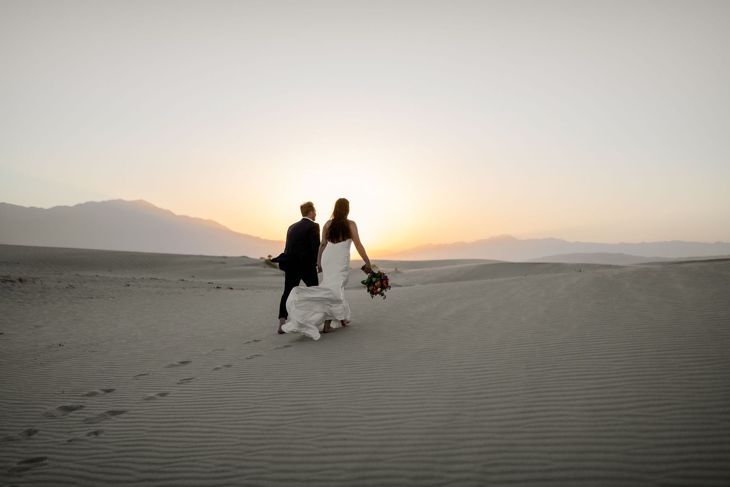 Bride and groom walk hand-in-hand across sand dunes at sunset in a stunning desert elopement.
