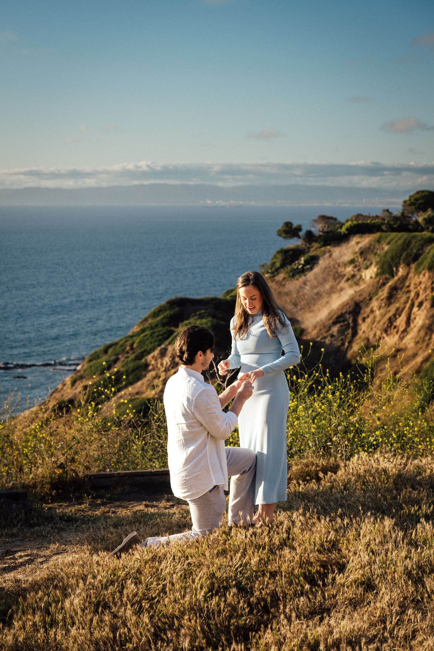 Man proposes on one knee to his partner on a scenic coastal cliffside overlooking the ocean at sunset.