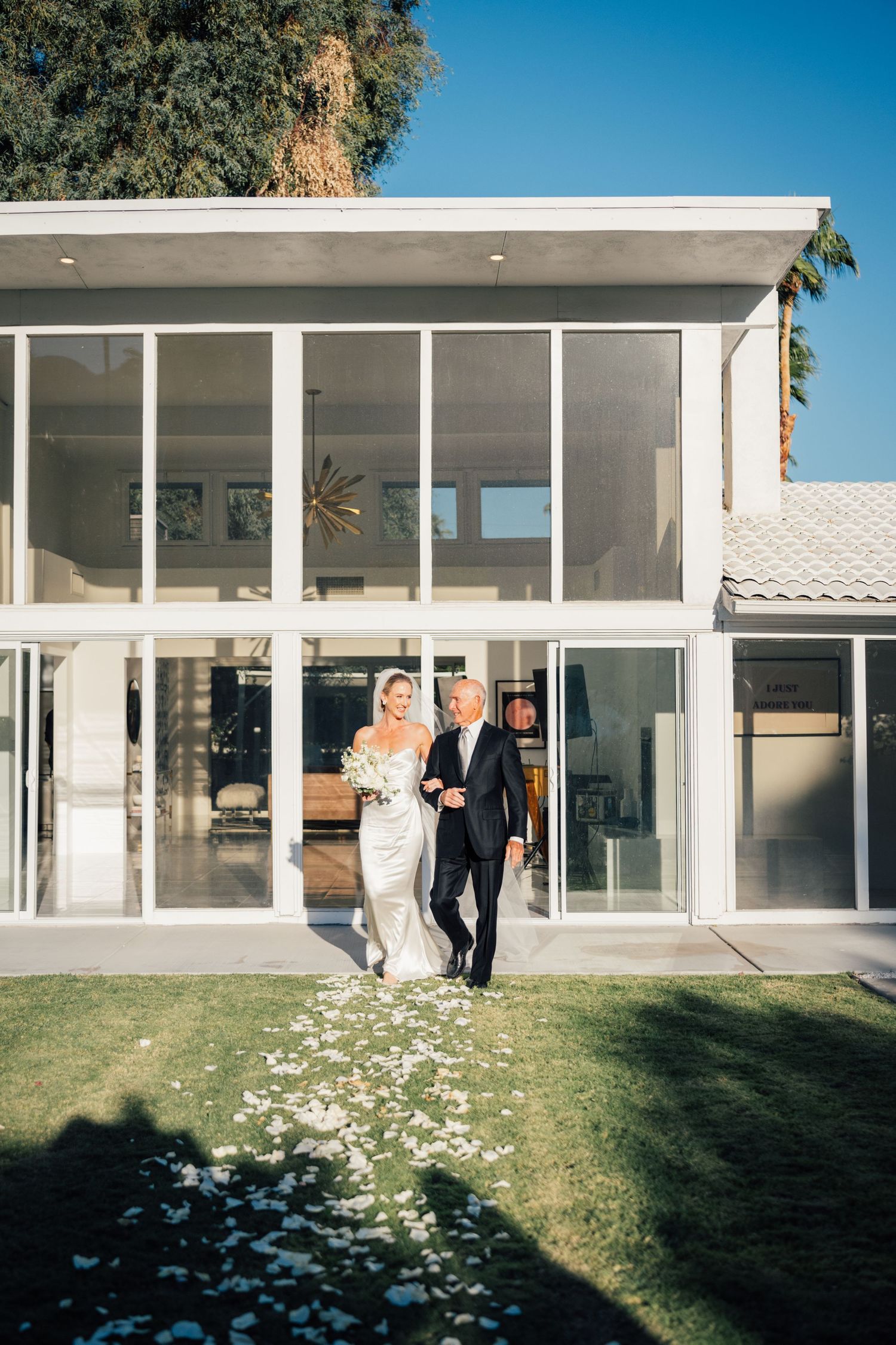 Louis Willard Photo Palm Springs Bride and father walk down a petal-lined aisle outside a modern glass-walled Palm Springs home.