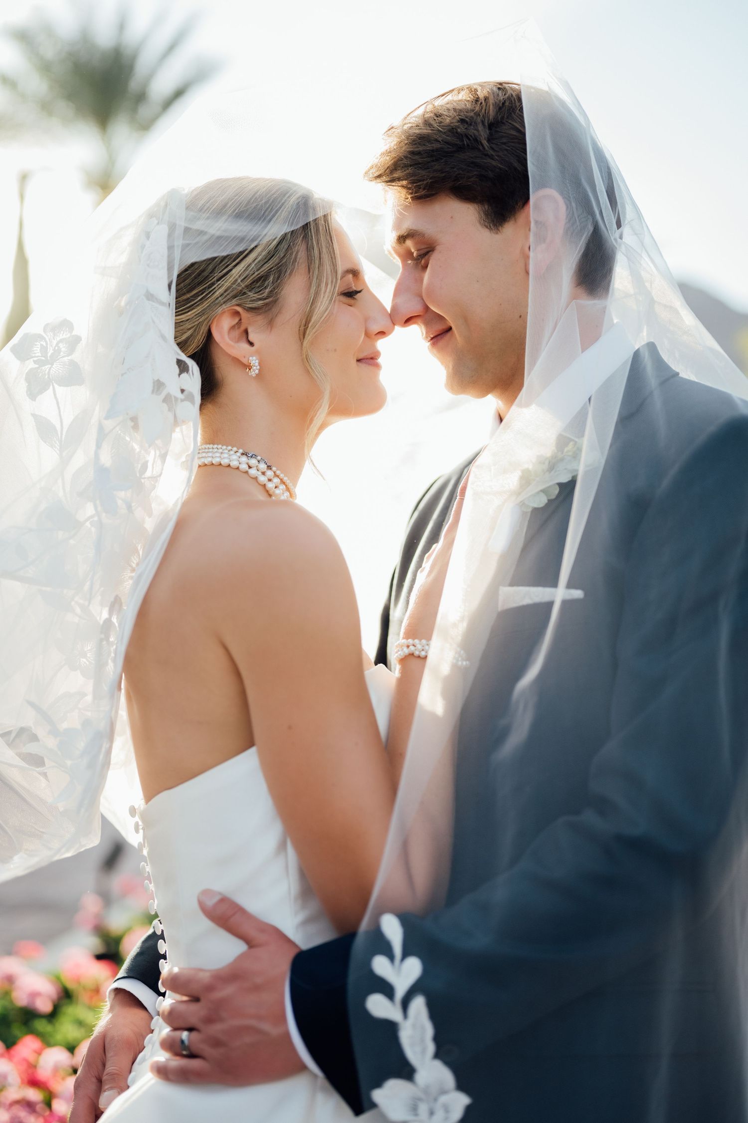 Bride and groom share romantic moment under flowing lace veil, nearly kissing outdoors on their wedding day.
