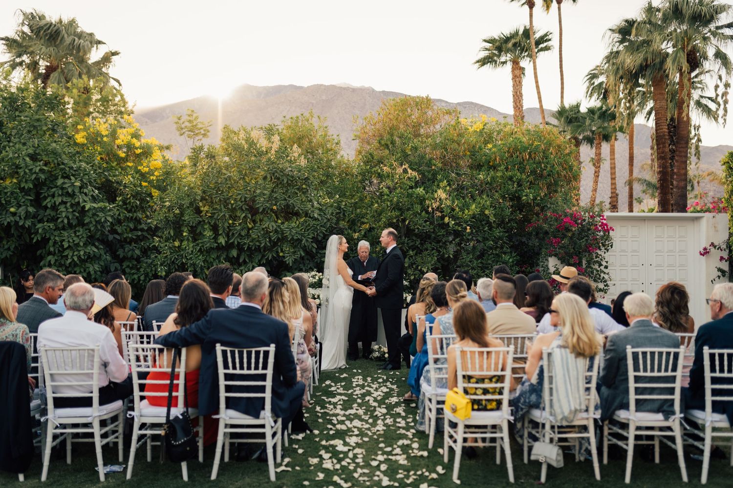 Louis Willard Photo Palm Springs Outdoor wedding ceremony with white chairs, floral aisle, palm trees, and mountain backdrop.