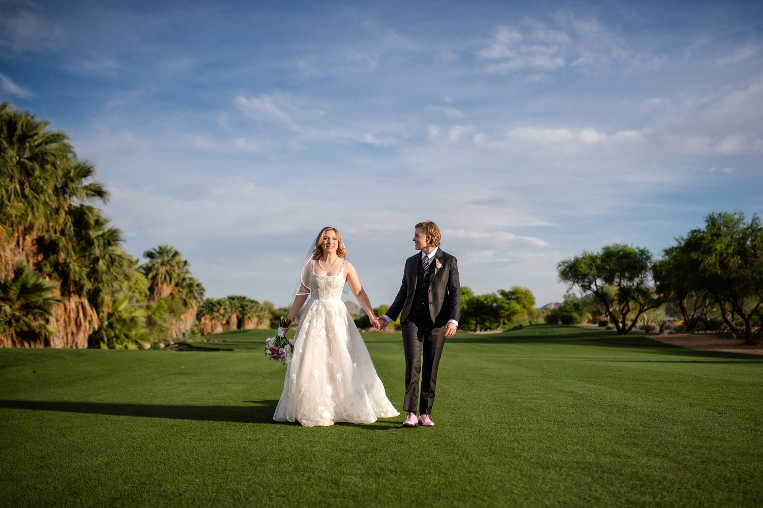 Bride and groom hold hands walking on lush green golf course with palm trees and blue sky at outdoor wedding.