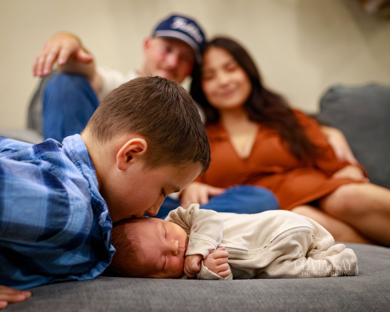 A young boy gently kisses a newborn baby on a couch while proud parents smile softly in the background.