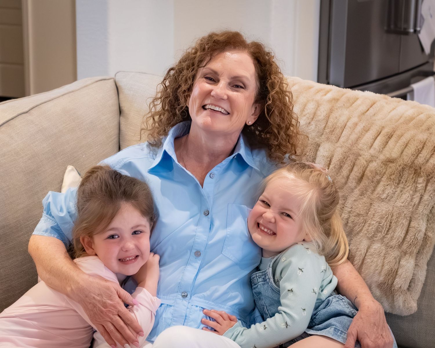 Woman with curly hair smiling on couch, hugging two young girls in a warm, cozy living room setting.