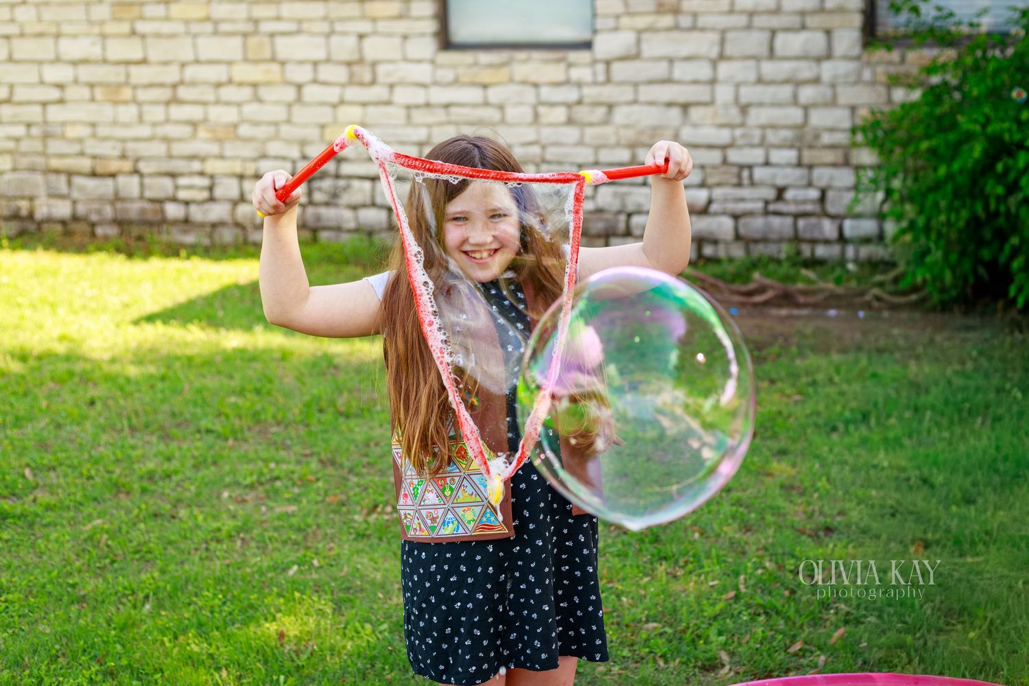 Young girl smiling while making a giant soap bubble with wands in a sunny backyard.