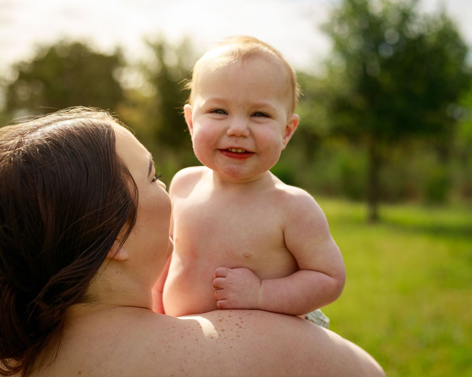 Happy baby smiling outdoors while being held by mother in sunny green park setting.