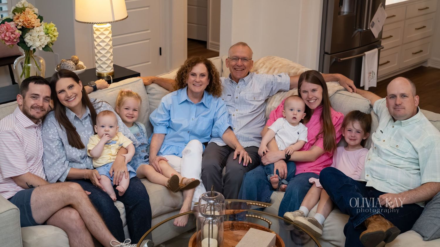 Large family group portrait with grandparents, parents, and young children seated together on a sofa in a cozy home.