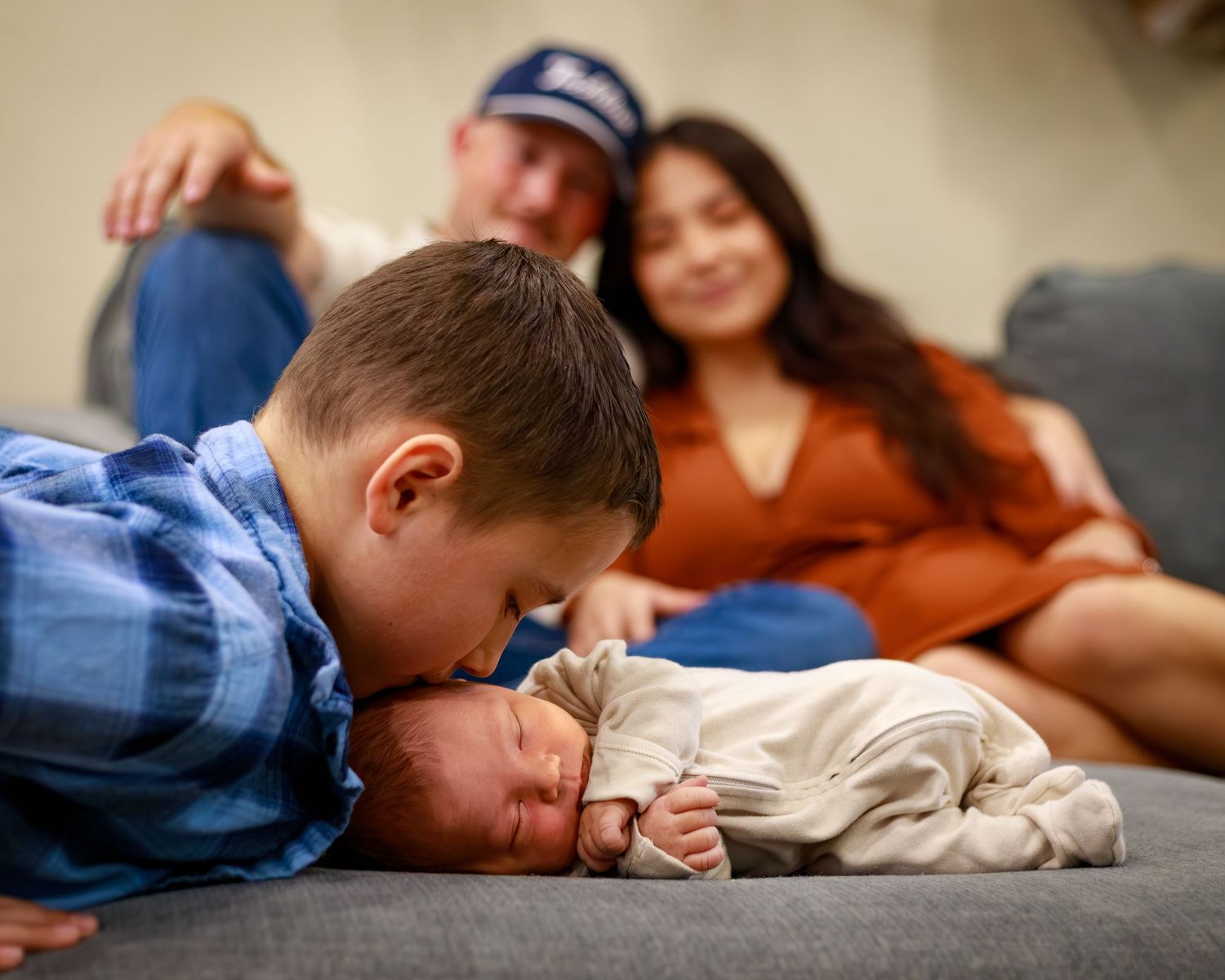 A young boy gently kisses a newborn baby on a couch while proud parents smile warmly in the background.