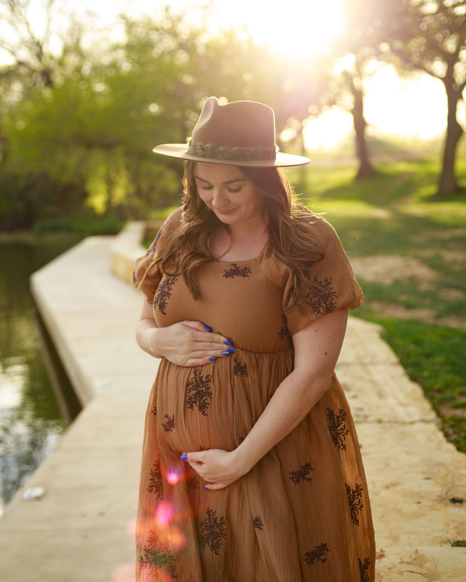 Pregnant woman in brown floral dress and wide-brim hat cradling baby bump at golden hour outdoors.