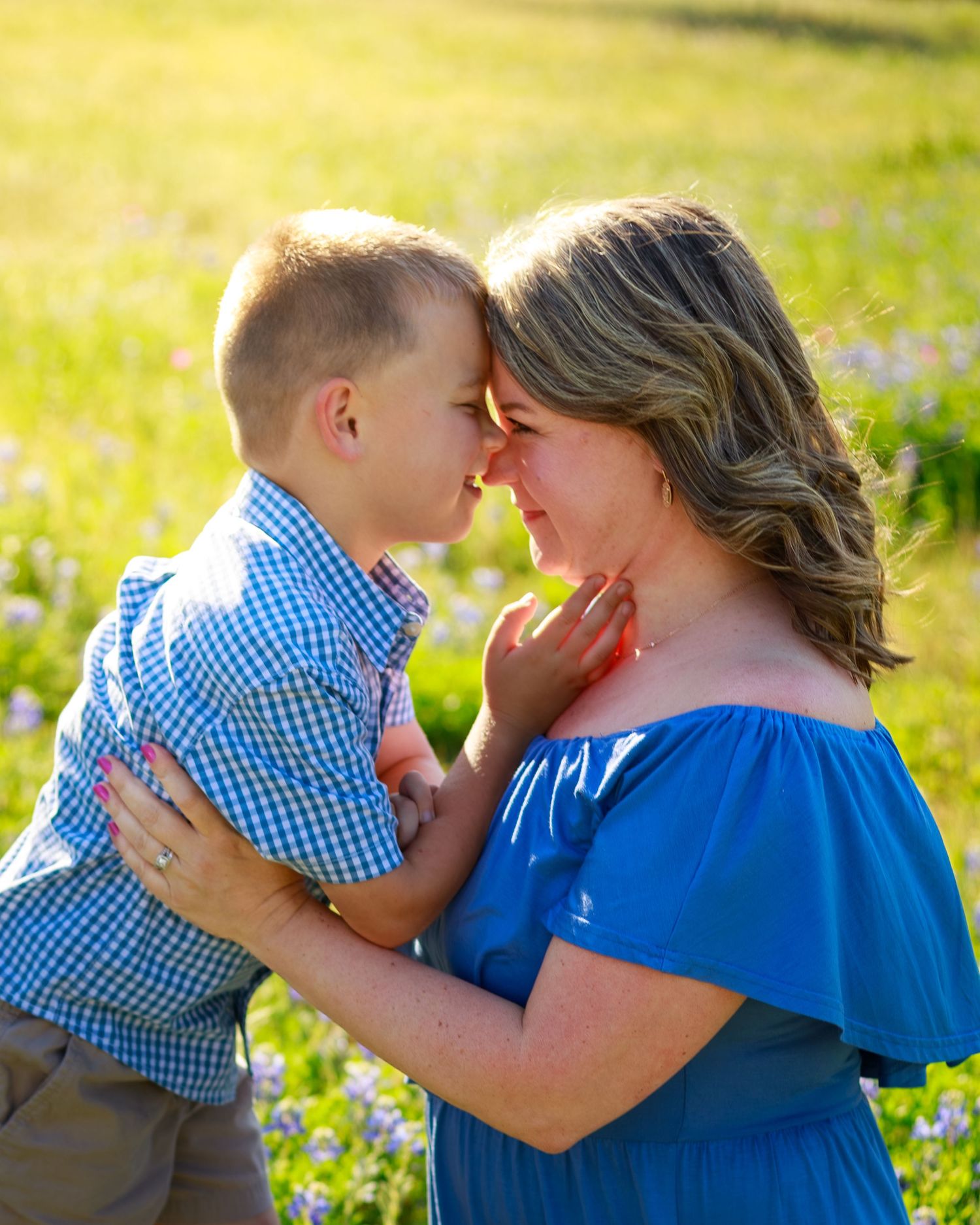 A mother and young boy share a tender nose-to-nose moment in a sunny green field with wildflowers.